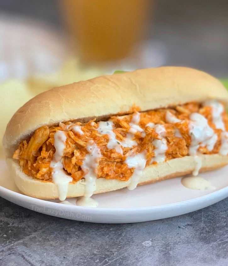 Buffalo chicken sandwich topped with creamy dressing on a white plate, ready to eat.